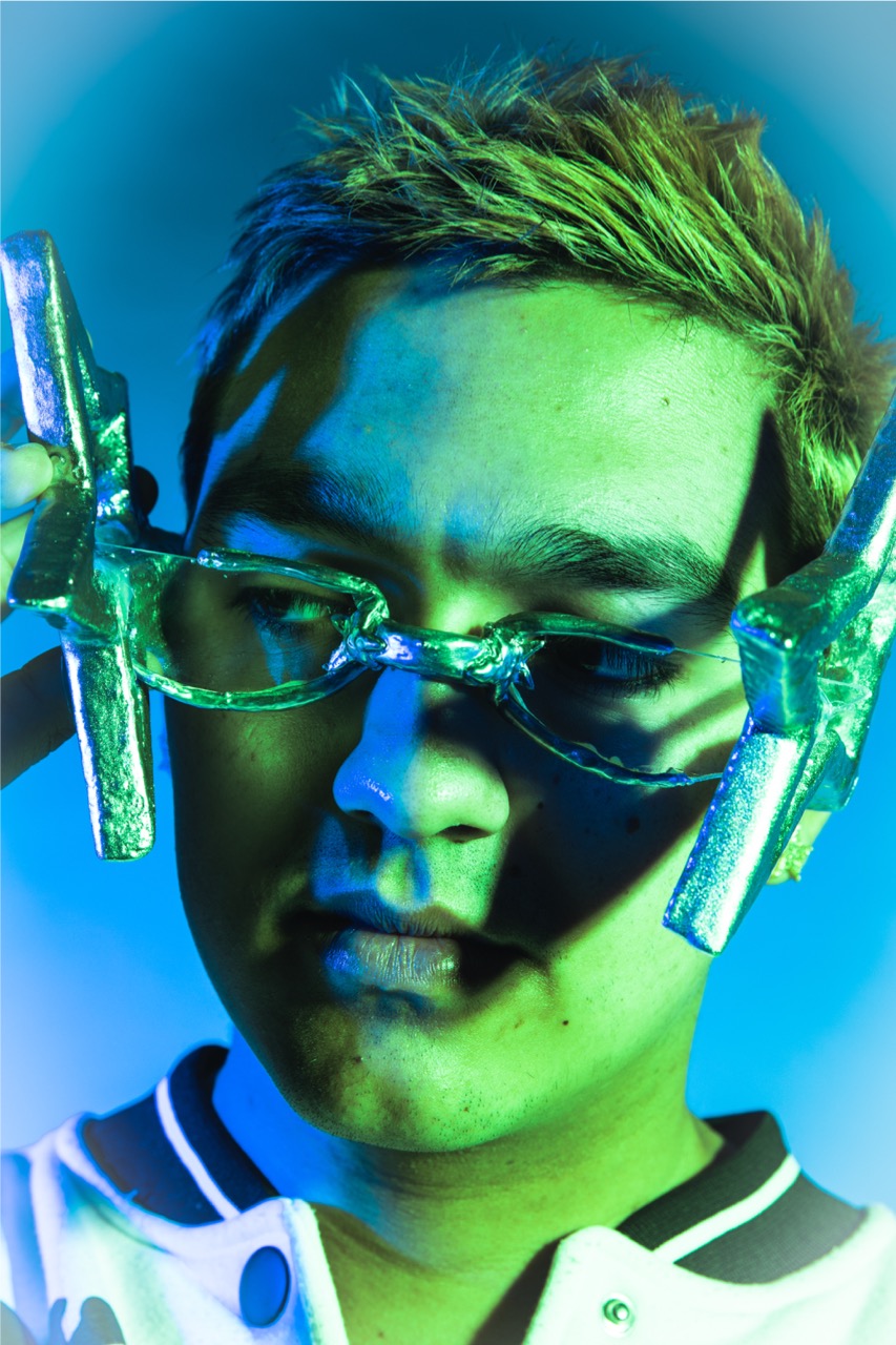 A close-up portrait of a young man wearing translucent glasses and a white jacket, lit by green and blue studio lighting against a plain background.