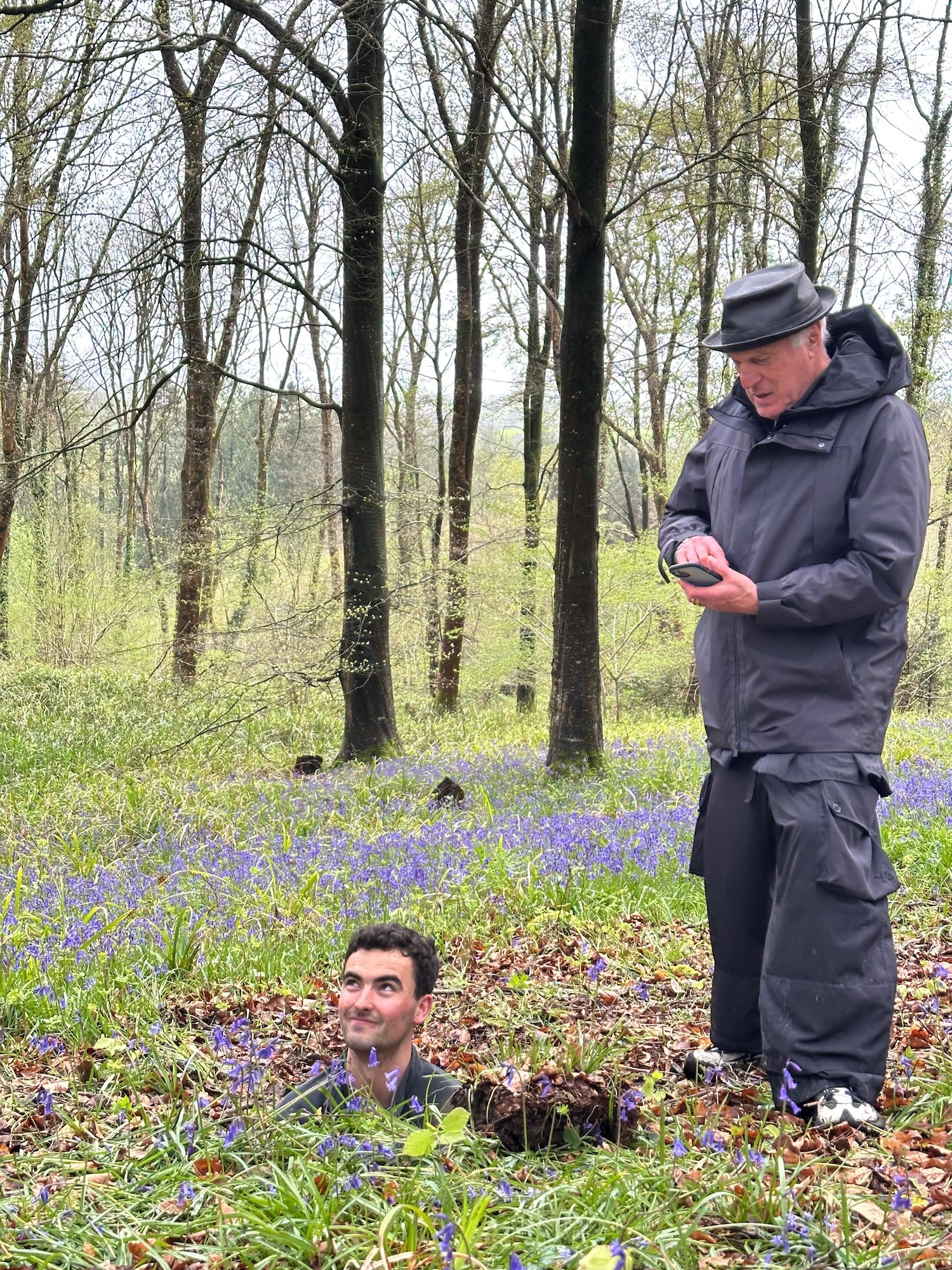 An older man in a grey jacket and hat stands in a woodland clearing of bluebells, while another man is partly buried in the ground in the foreground.