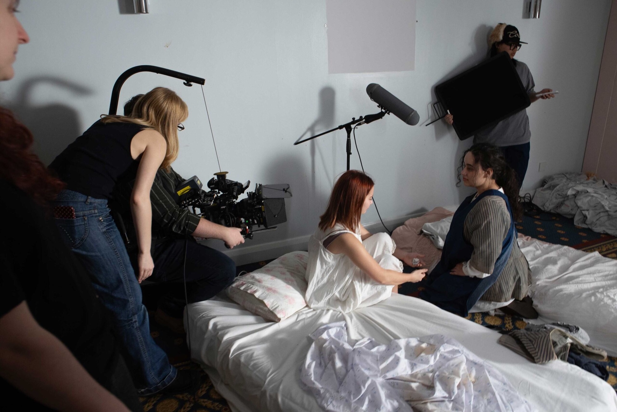 A film crew records two women seated on a bed in a sparsely furnished bedroom, with a camera rig and boom microphone in the foreground.