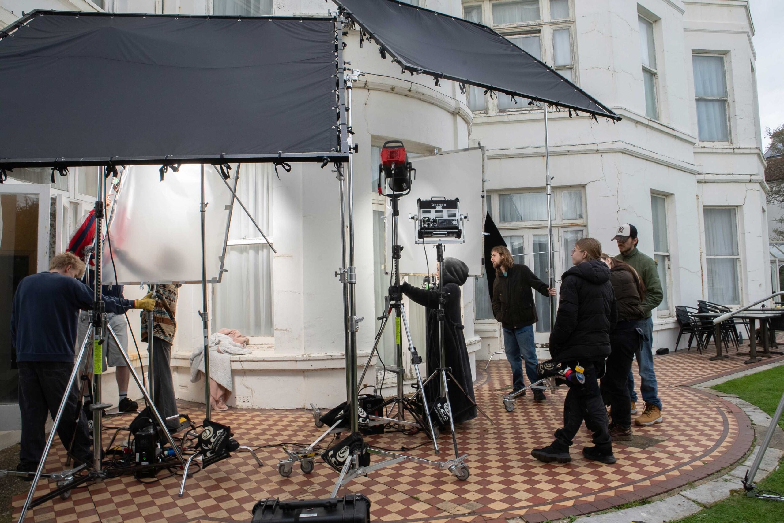 A film crew operates on location outside a white building. Various lighting equipment and camera rigs are set up, while crew members adjust settings and discuss production. The scene is set on a patterned outdoor floor, with furniture visible in the background.