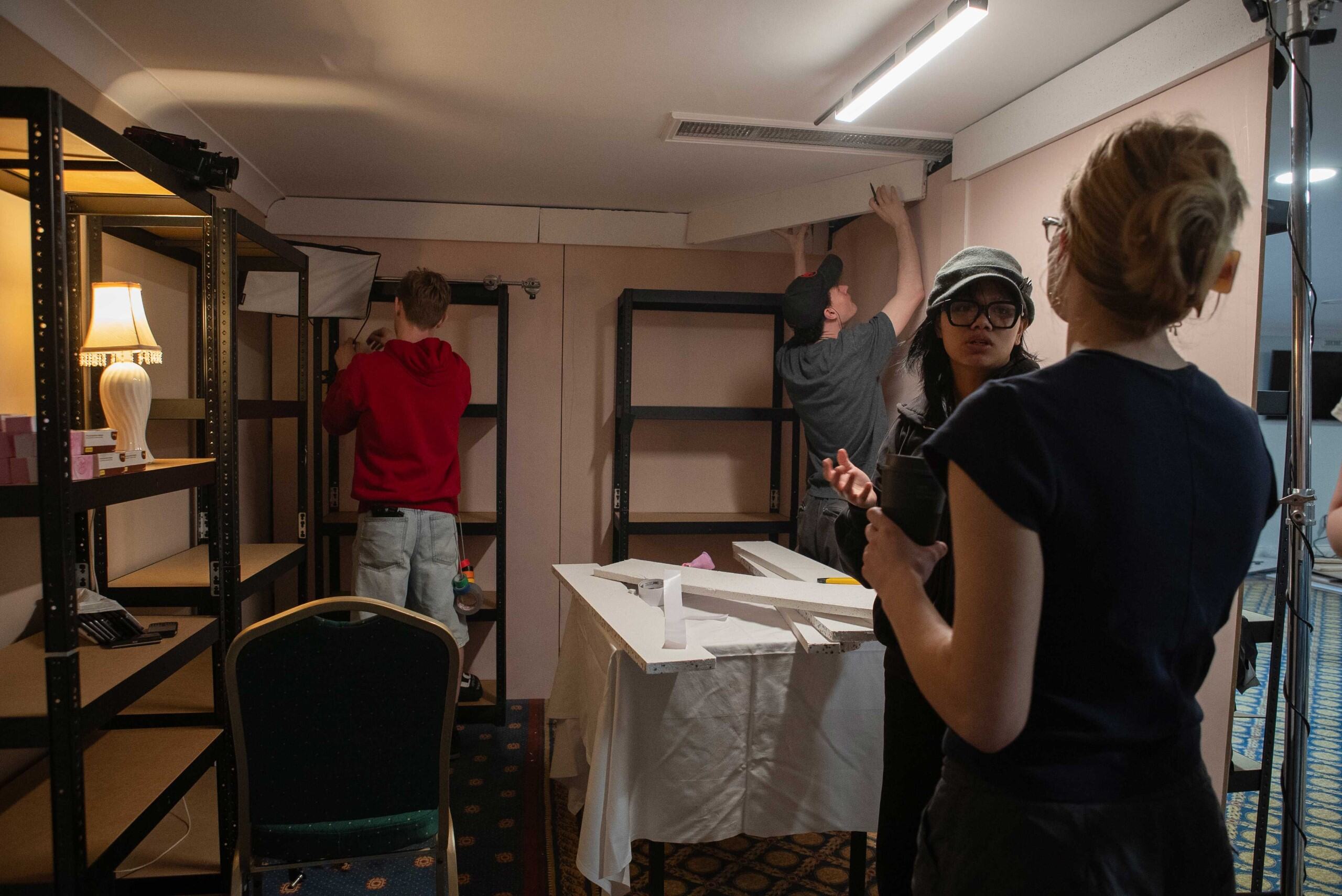 A group of three individuals is working on setting up a space with shelves. Two people are adjusting a light fixture, while another is holding documents and standing next to a table covered with a white cloth. There are empty shelves and a lamp in the background.