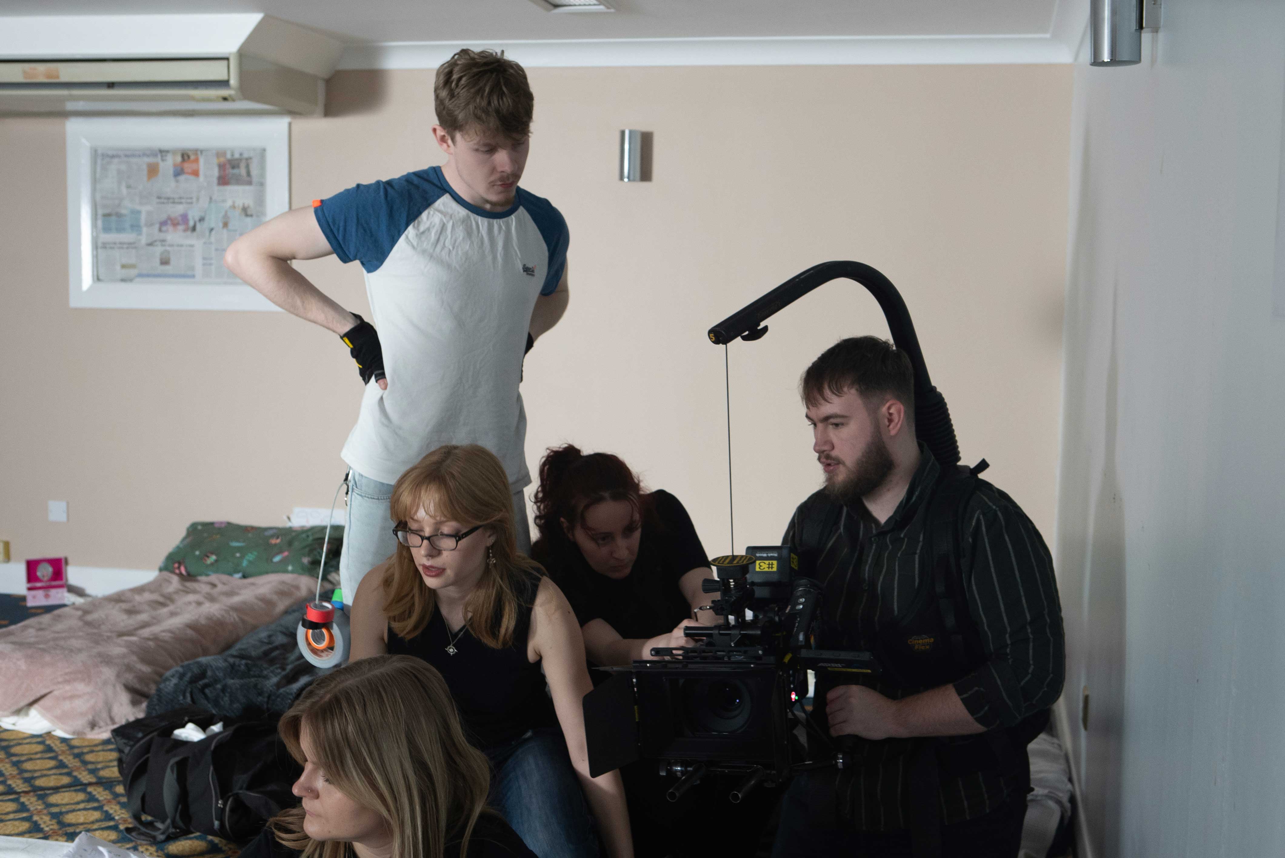Five individuals are engaged in a film shoot in a room with soft furnishings. One man stands with his hands on his hips, while another adjusts a camera rig held by a bearded man seated. A woman with glasses watches attentively.