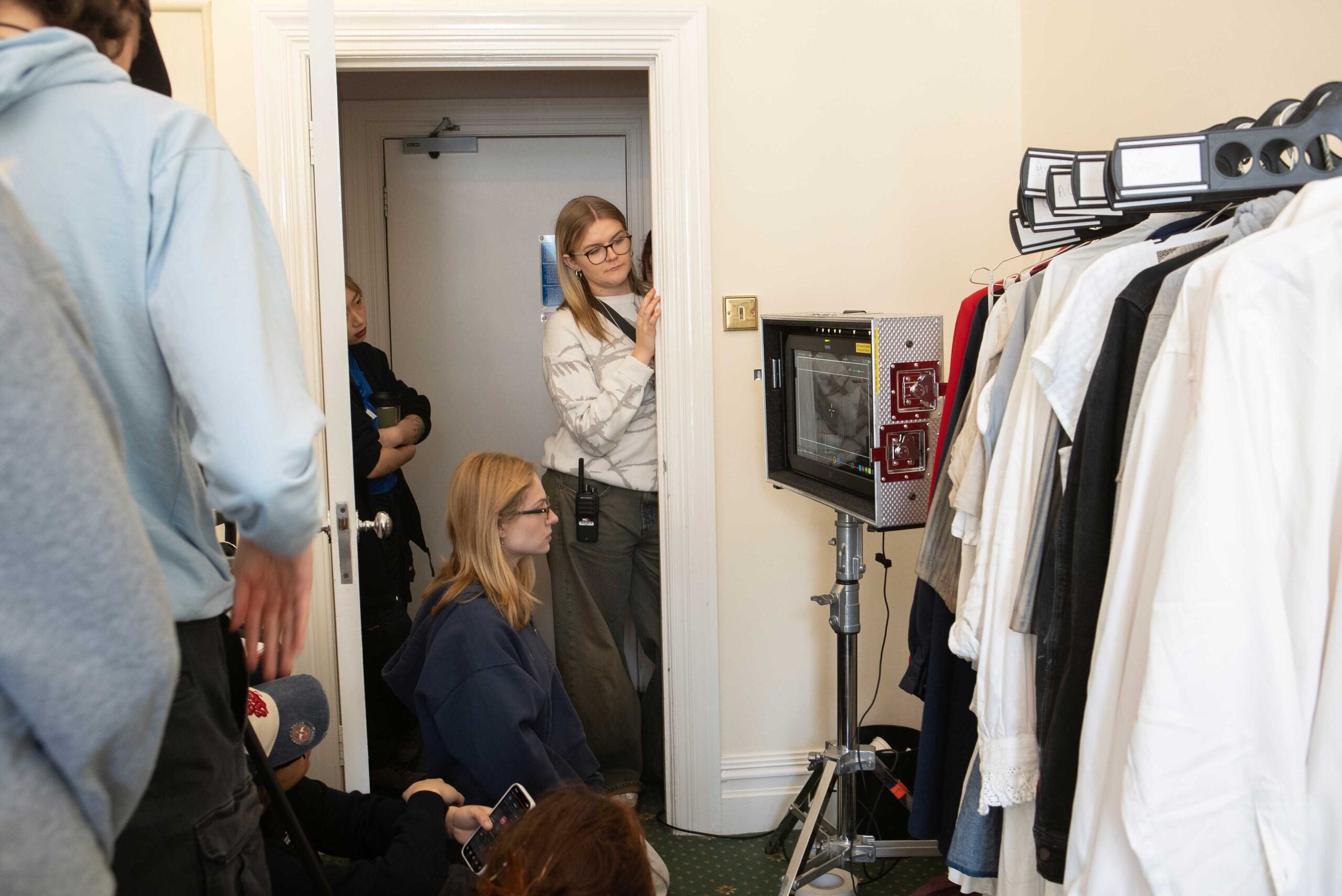 A group of people stands near a doorway, with one woman in a white top observing a monitor displaying a fashion show. In the foreground, a rack holds various garments and the room features light-coloured walls.