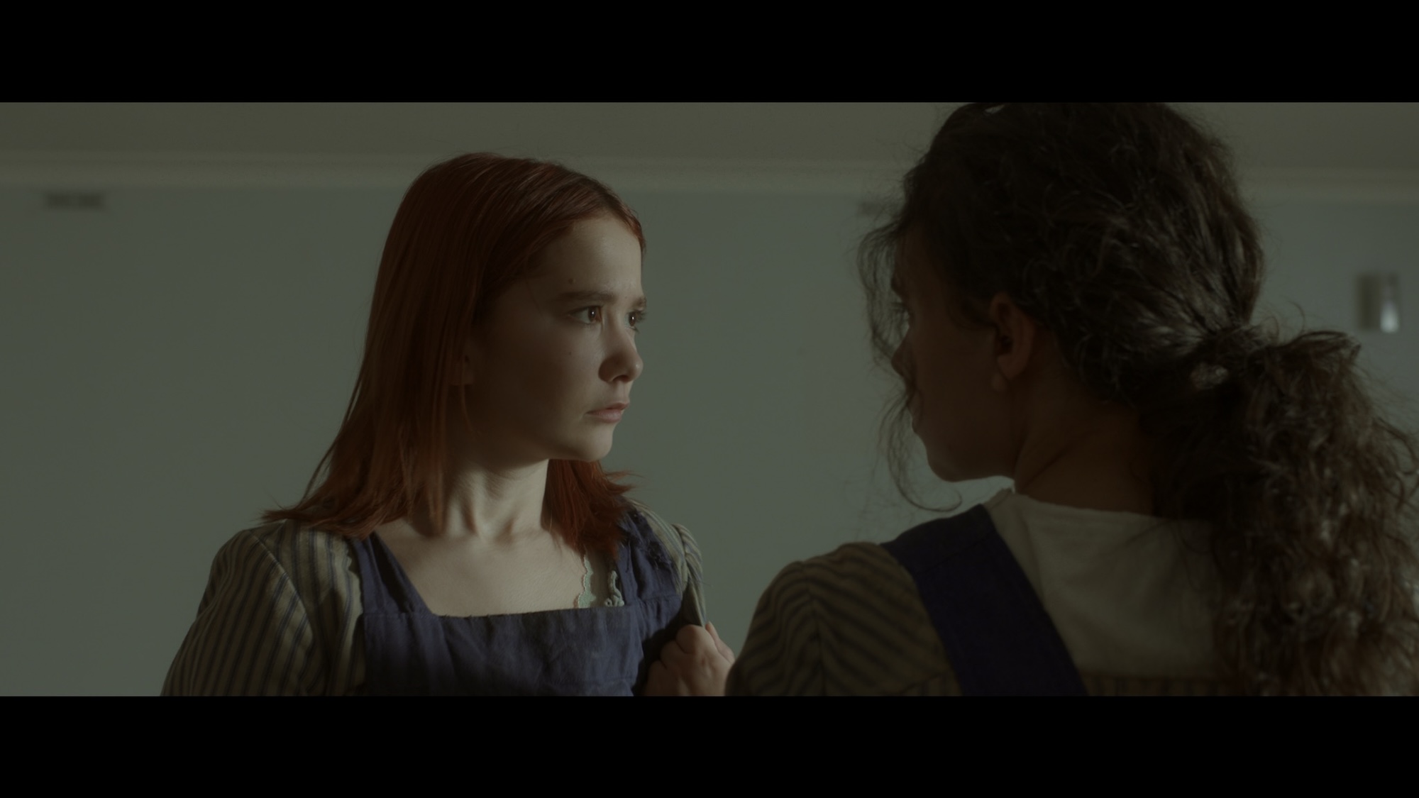 Two young women stand closely together in a dimly lit room. The woman on the left has reddish-brown hair and wears a blue dress with white stripes, while the woman on the right has dark curly hair and is dressed in a similarly styled outfit.