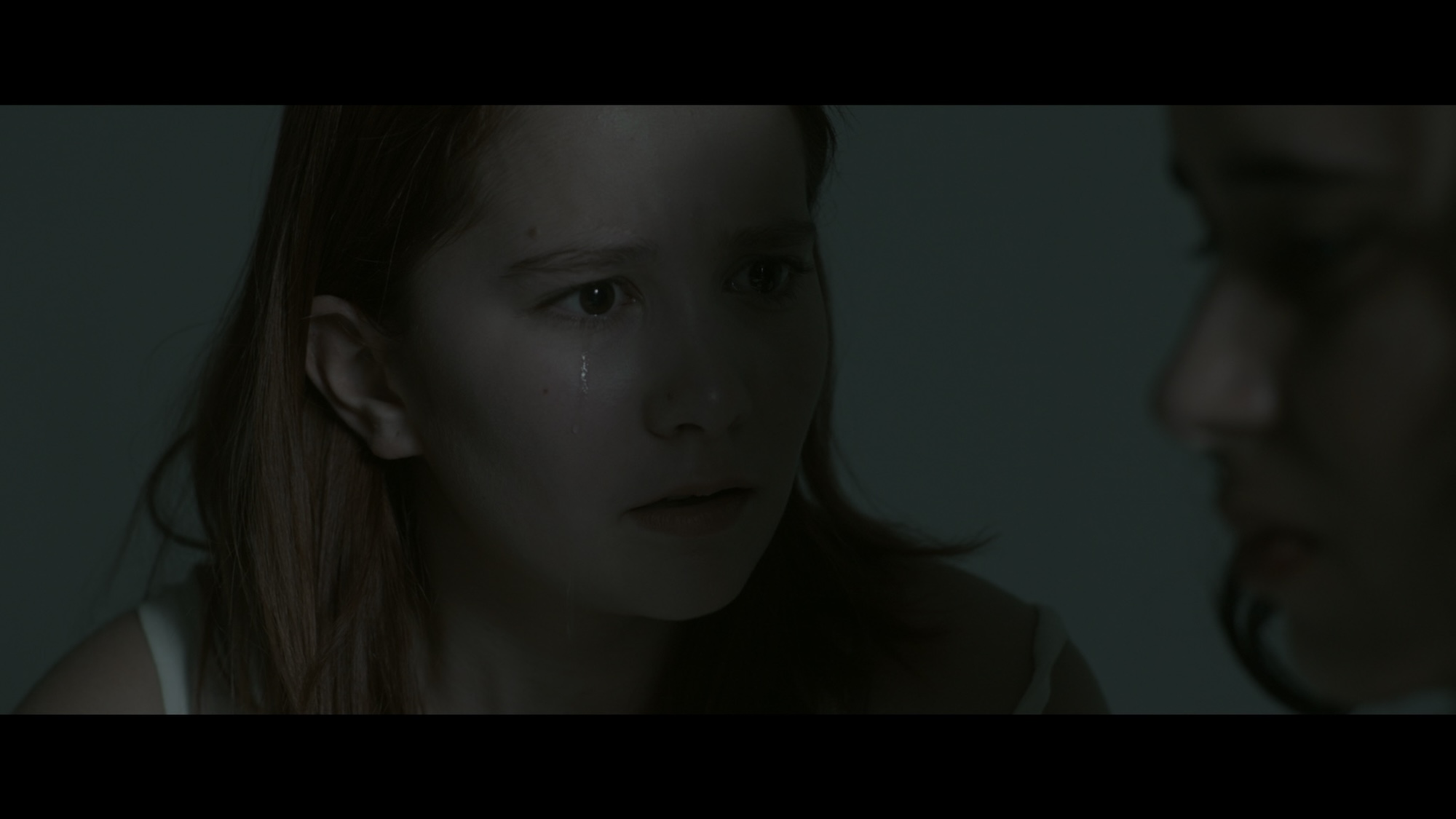 A girl with long reddish hair gazes intently, with a tear rolling down her cheek, while sitting close to another girl out of focus in the foreground. The dim lighting creates a muted atmosphere in the background.