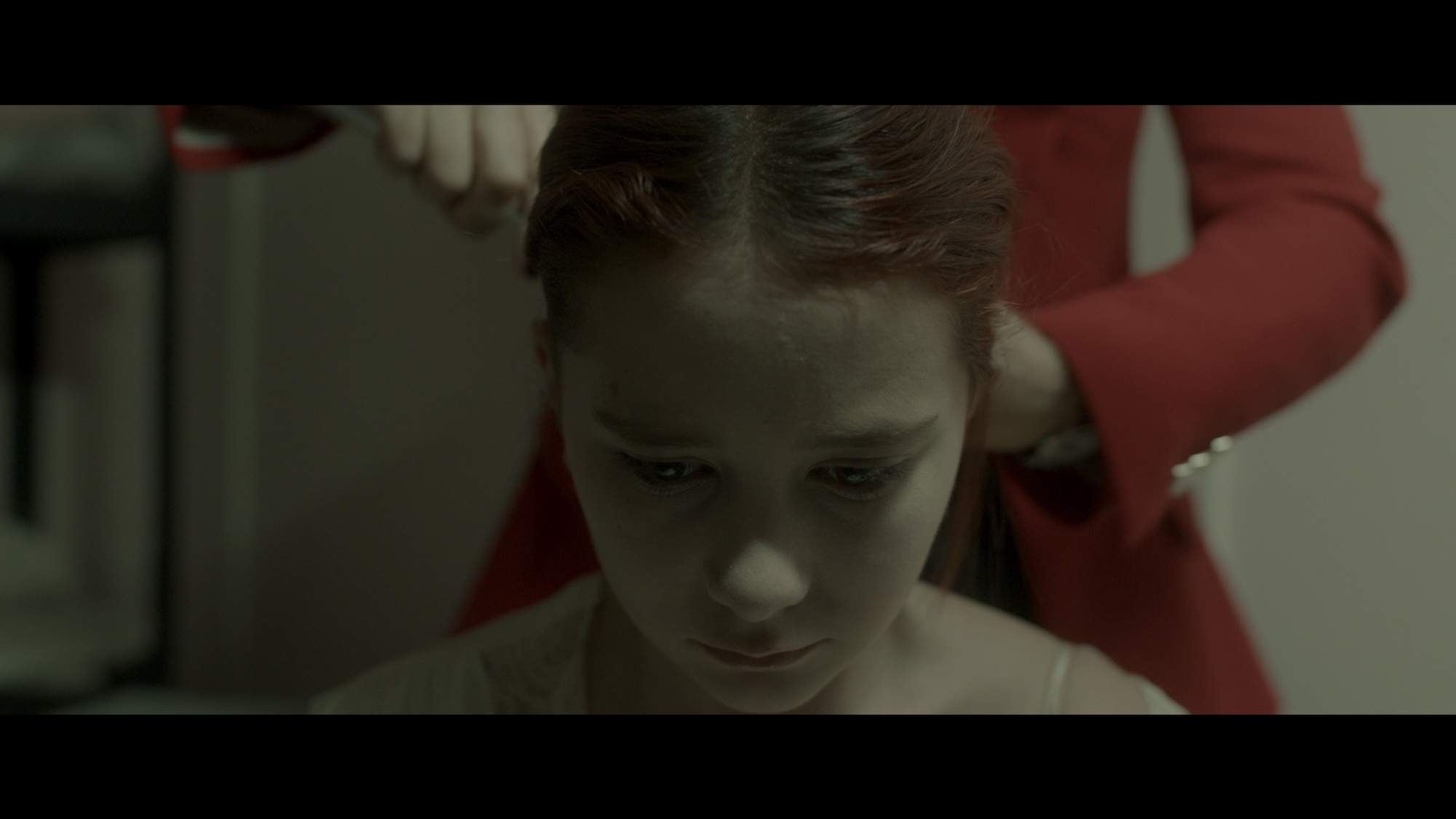 A young girl with brown hair is sitting down, looking downwards. A person in a red jacket is seen behind her, brushing the girl's hair. The background is softly blurred, suggesting a domestic setting.