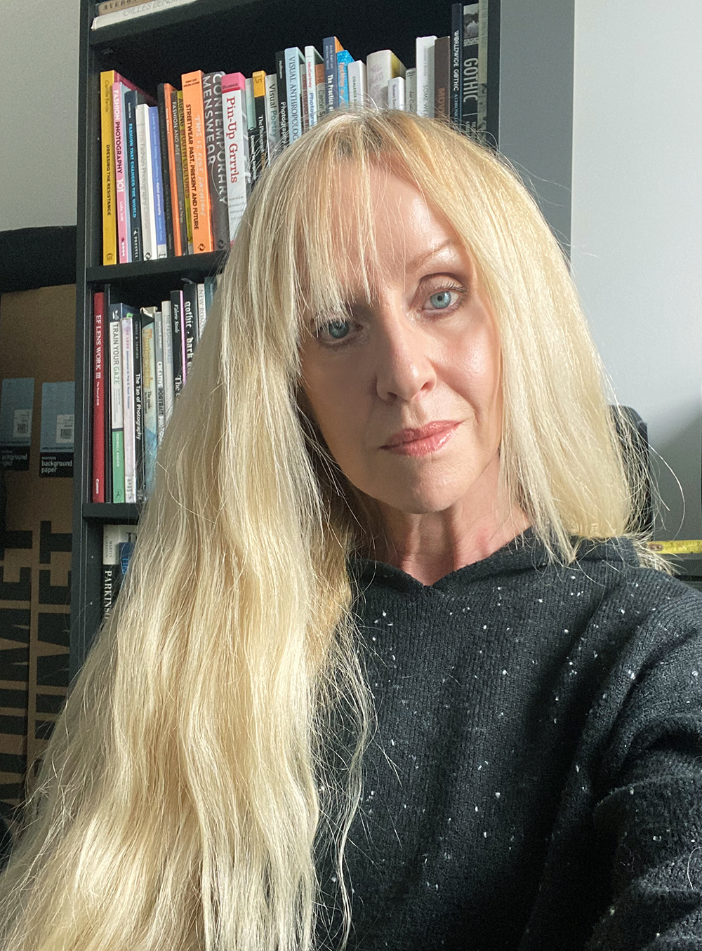Blonde woman in dark sparkly top sits in front of bookshelf filled with colourful books on black shelves.