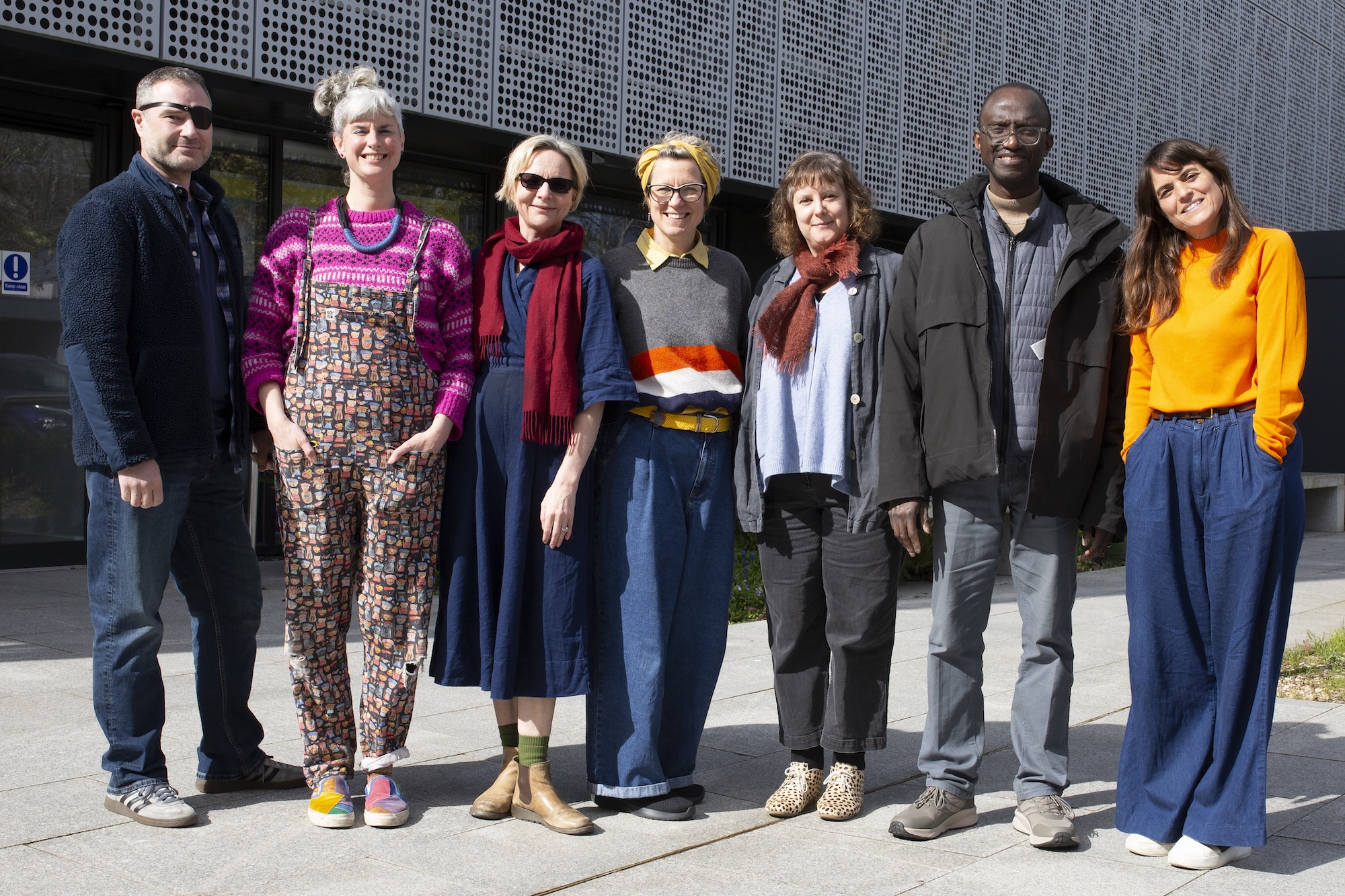 Seven adults stand together outdoors beside a modern building with a perforated metal facade.