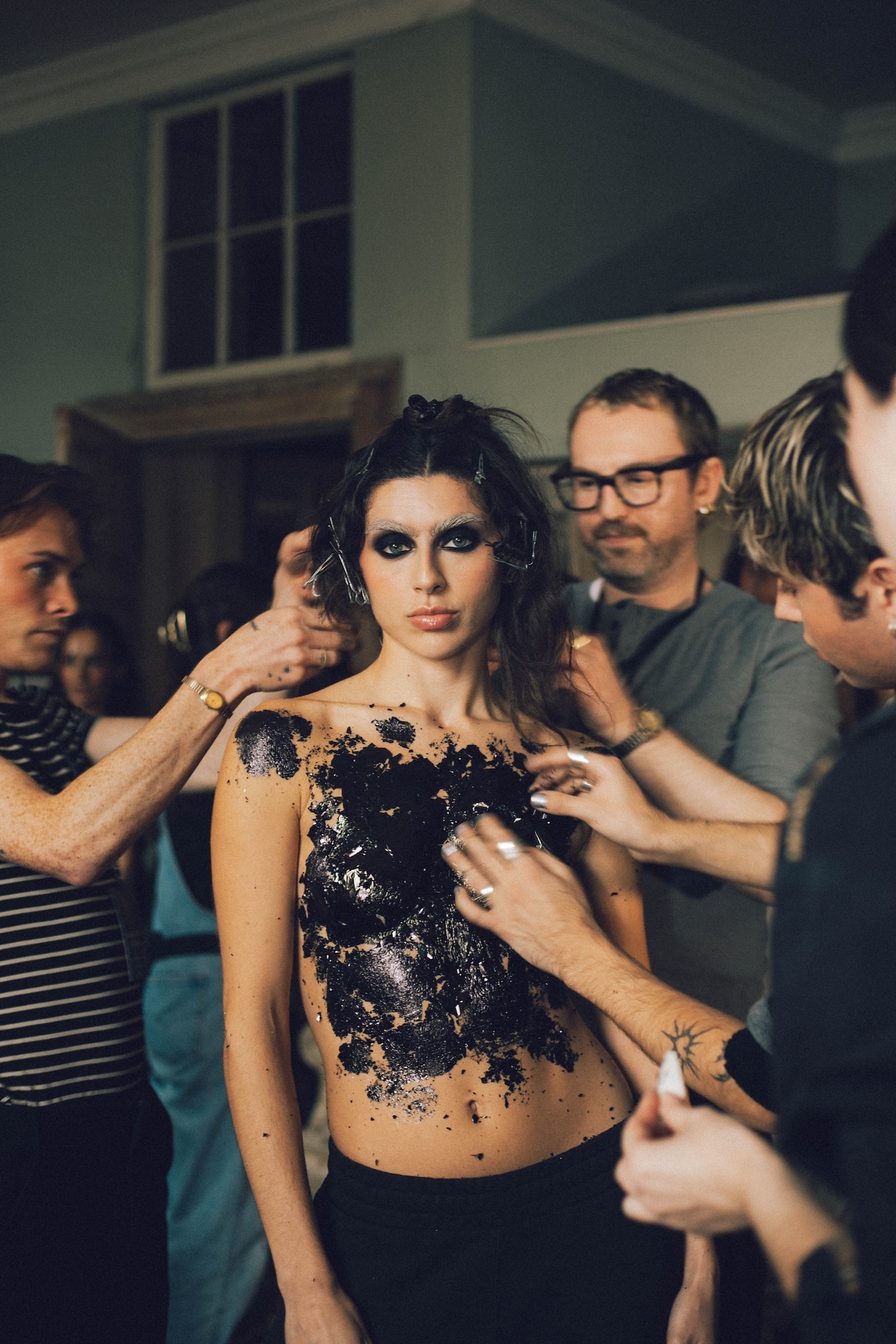 Woman with dark eye makeup and sequined top being styled by multiple people, man with glasses in background, indoor setting