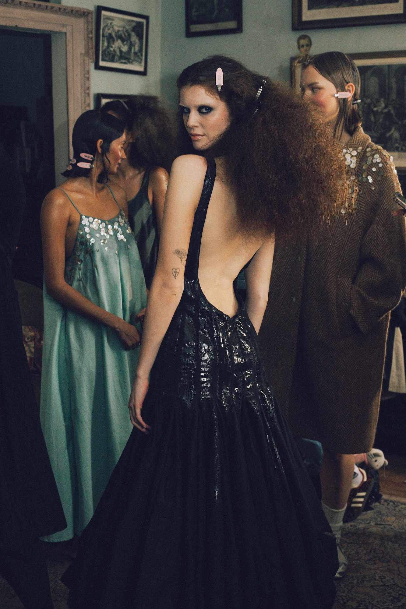 Three women in evening gowns - one in mint green, one in black sequined dress, one in gold sequined dress - standing in room with framed artwork