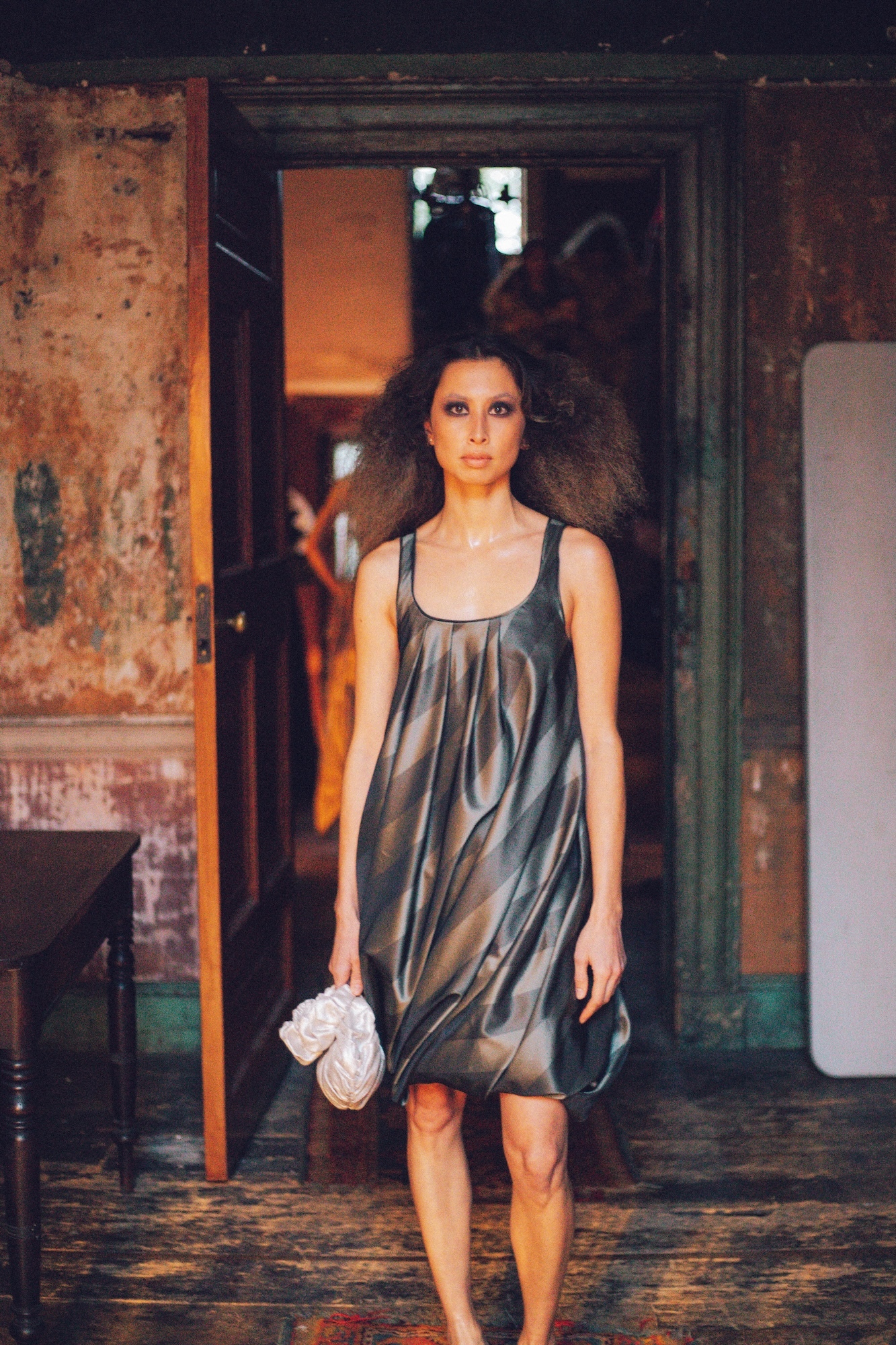 Woman in metallic grey dress holding white clutch bag, standing in doorway of weathered building with peeling paint