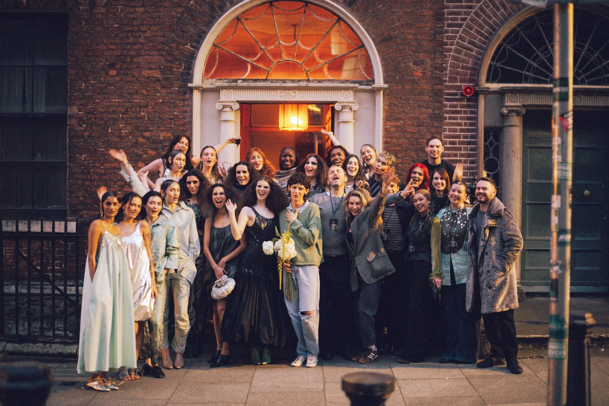 Large group of people posing together outside red brick building with arched doorway and warm lighting from entrance