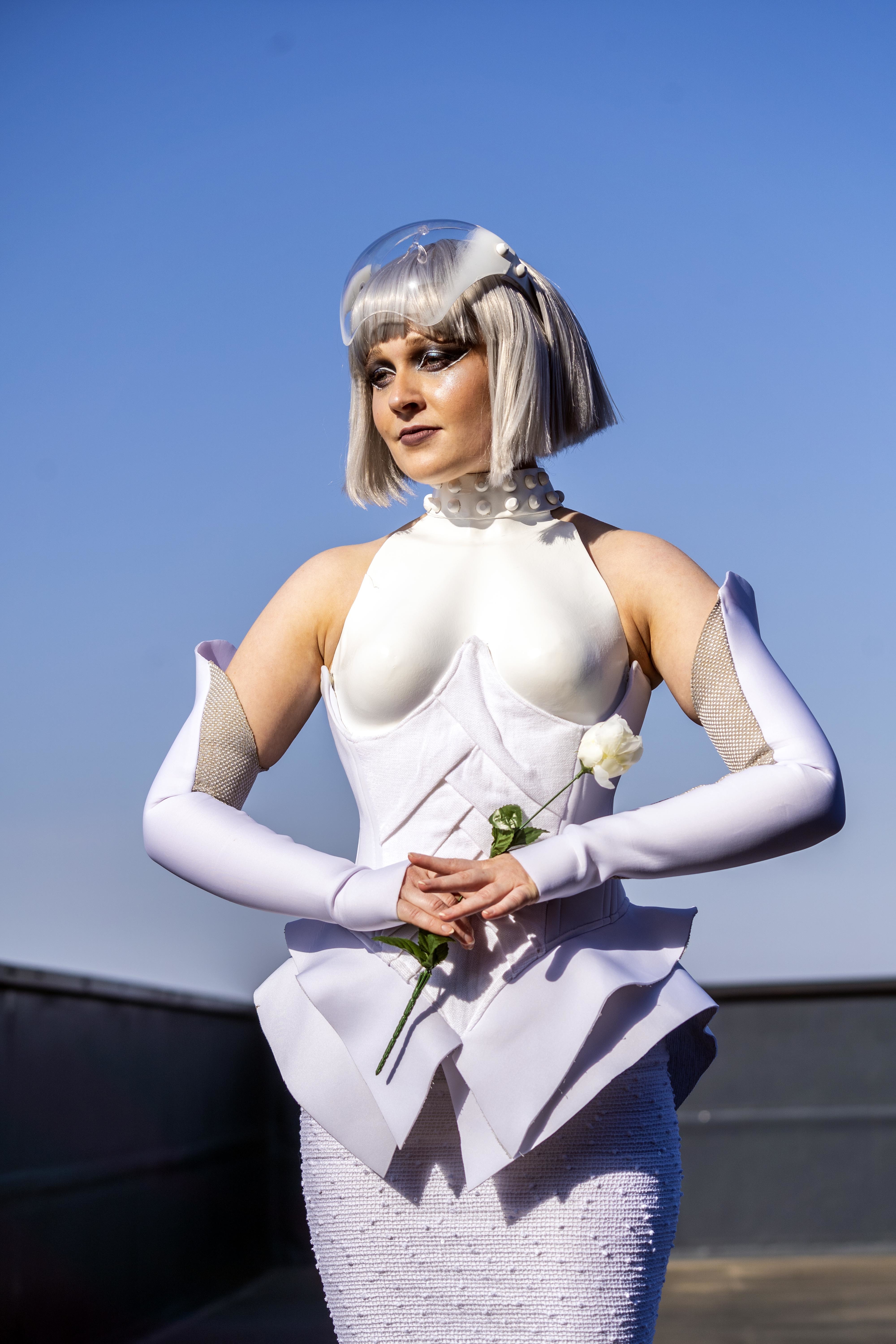 A model dressed in a Hunger Games-inspired costume stands holding a rose, with a clear blue sky behind them.