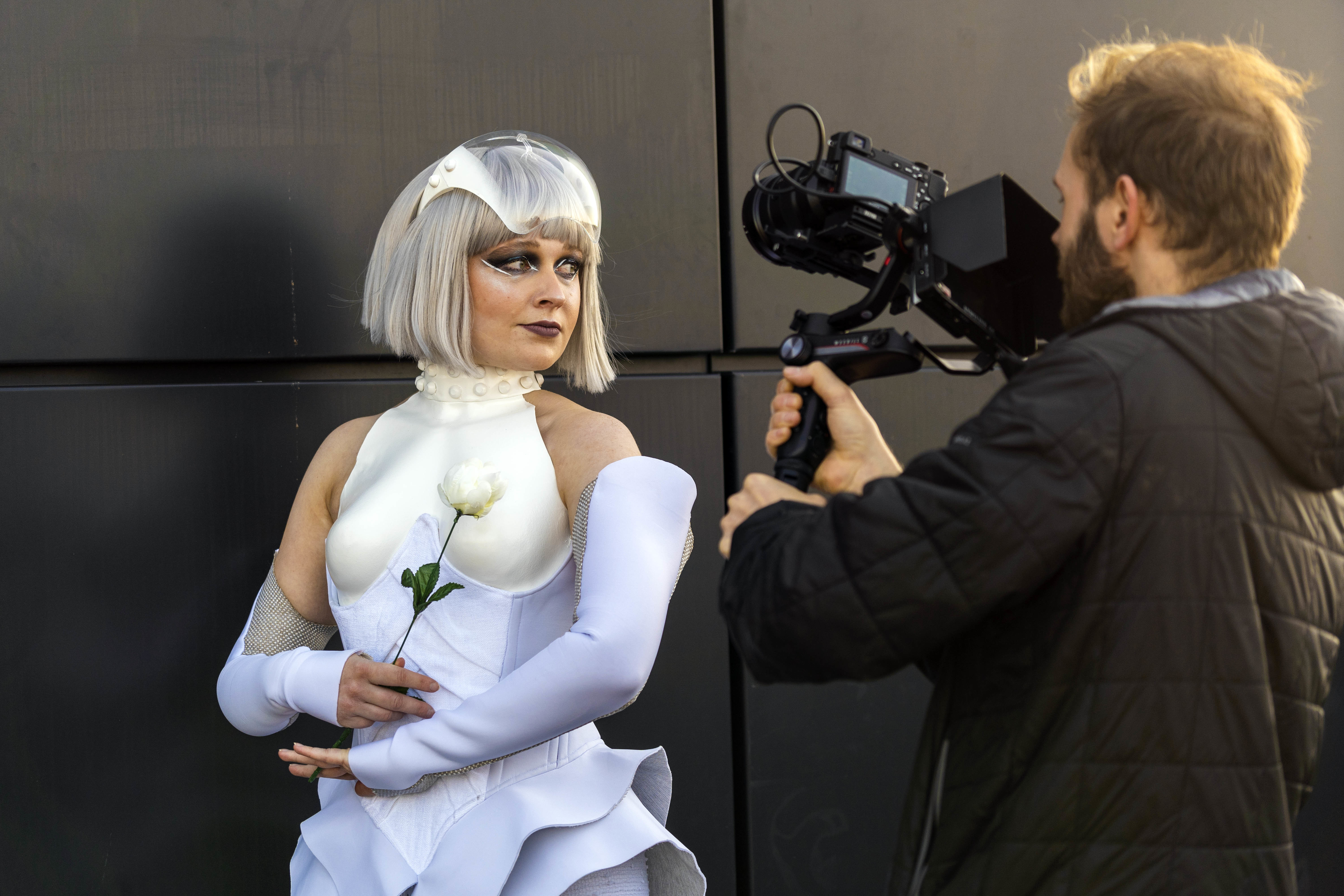 A model in a Hunger Games-inspired costume holds a rose and poses for a photo in front of a cameraman holding a camera.