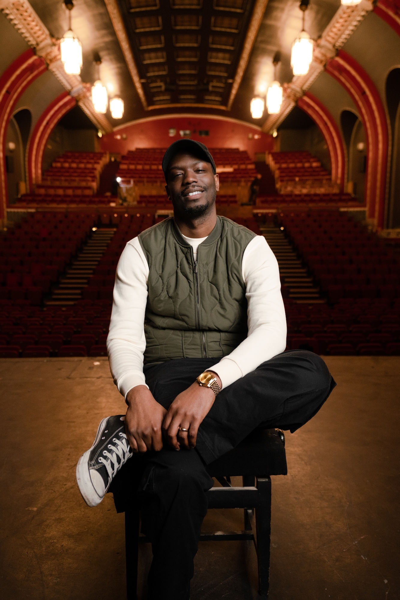 Playwright and actor R Calais Cameron sitting on a stage with an auditorium seating behind him.