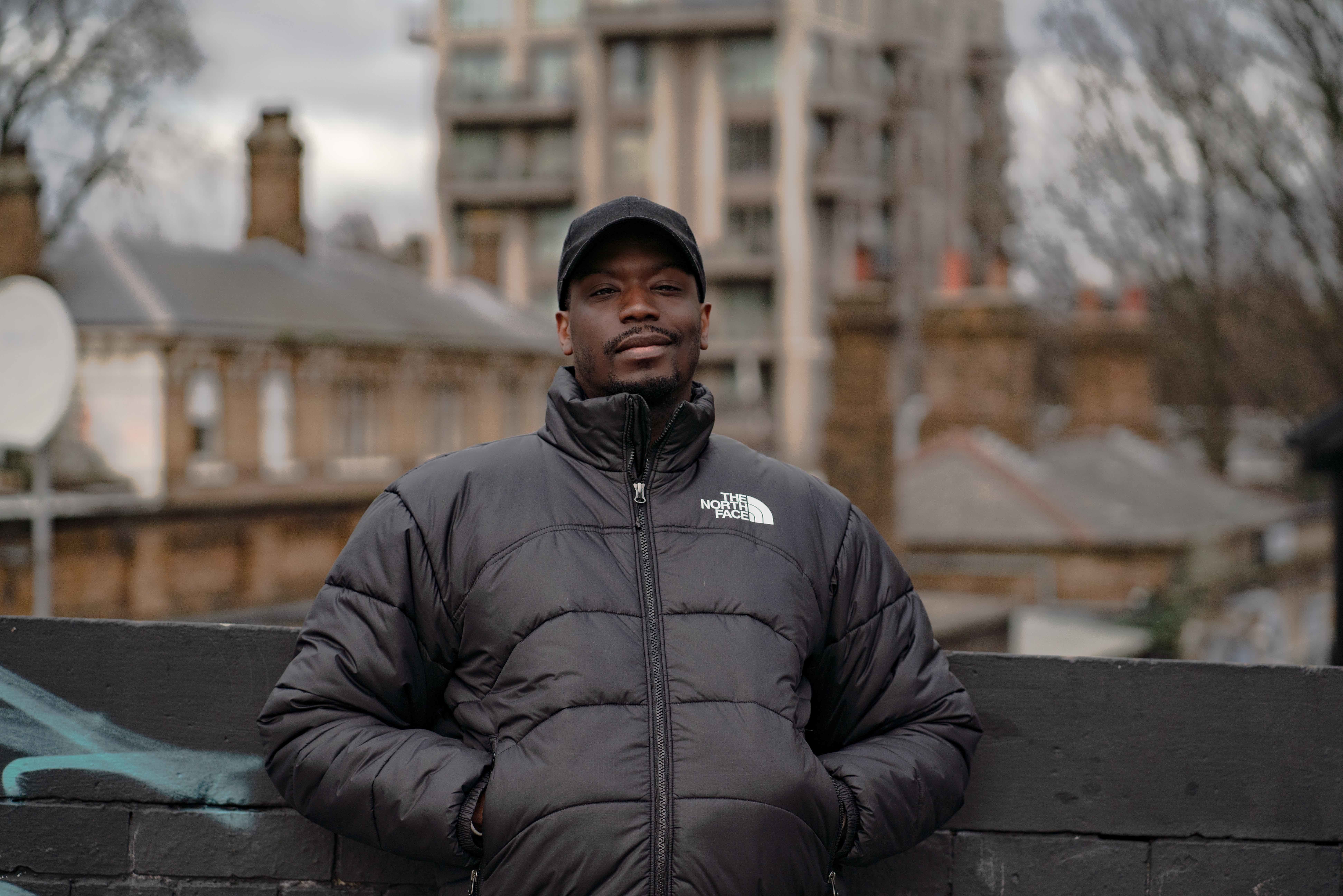 Headshot for R Calais Cameron, leaning up against a brick wall with city buildings in the background.