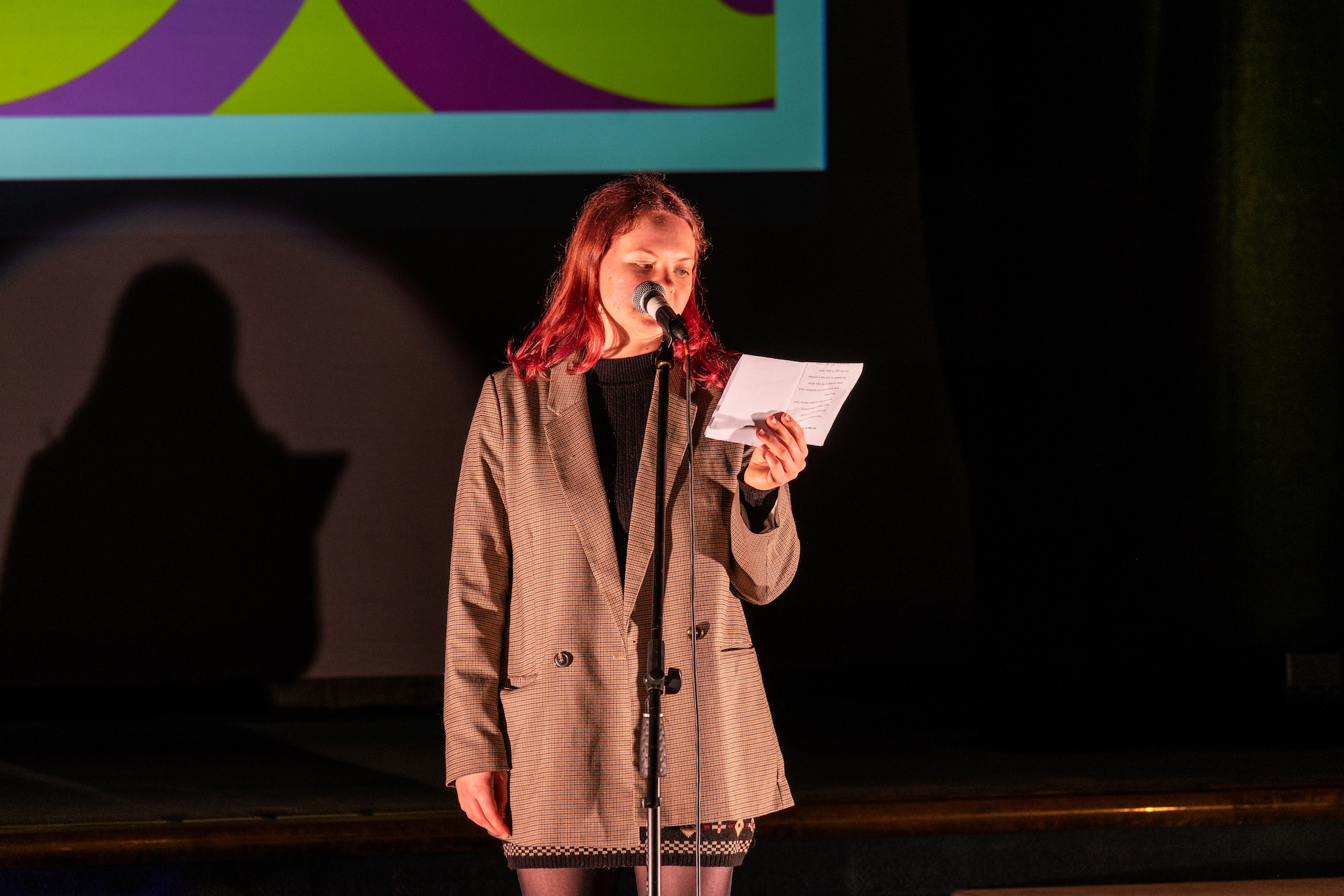 A student stands by a microphone on stage and reads from a piece of paper.