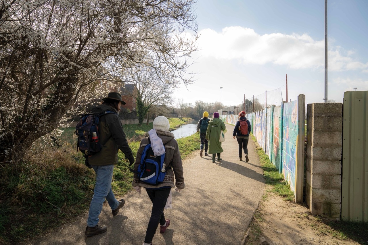 People walking down a path by a river and trees, sunny day.