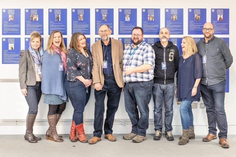 A group of eight people stands and poses for a photo in front of a wall of framed posters
