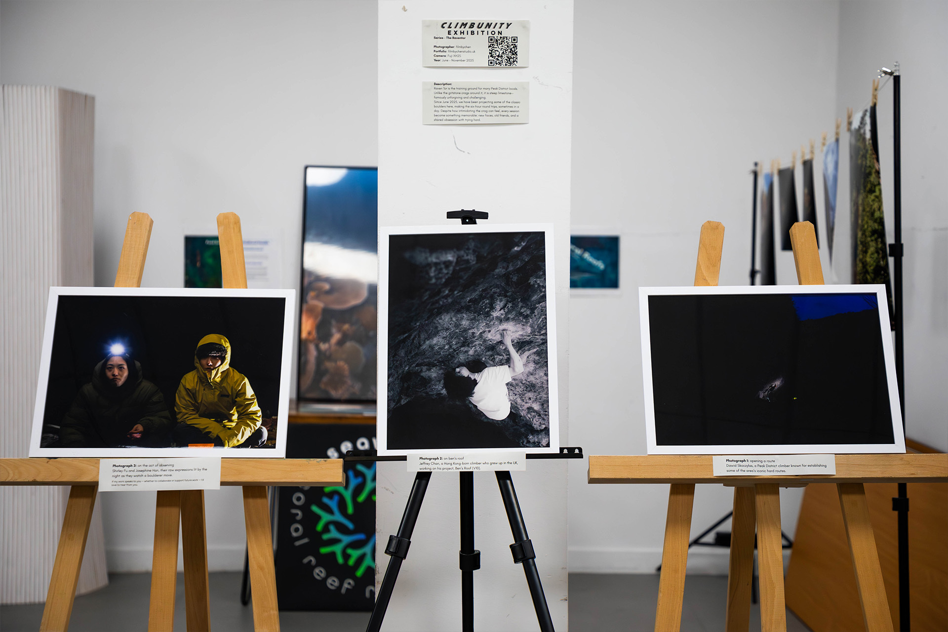 Photographs displayed on wooden easels in white gallery space, including images of people and landscapes, with wall-mounted artwork behind.