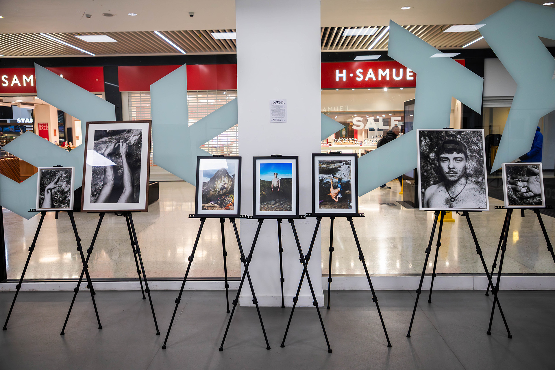 Gallery display of framed photographs on black tripod easels in shopping centre with H Samuel store visible in background.