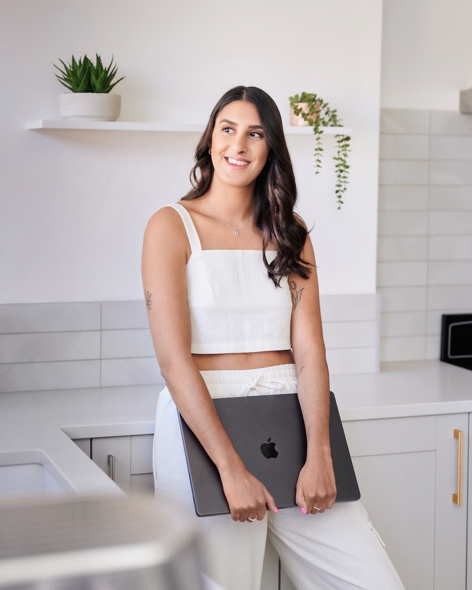 T Johal holding laptop in modern kitchen with white tiles, floating shelves, and potted plants.