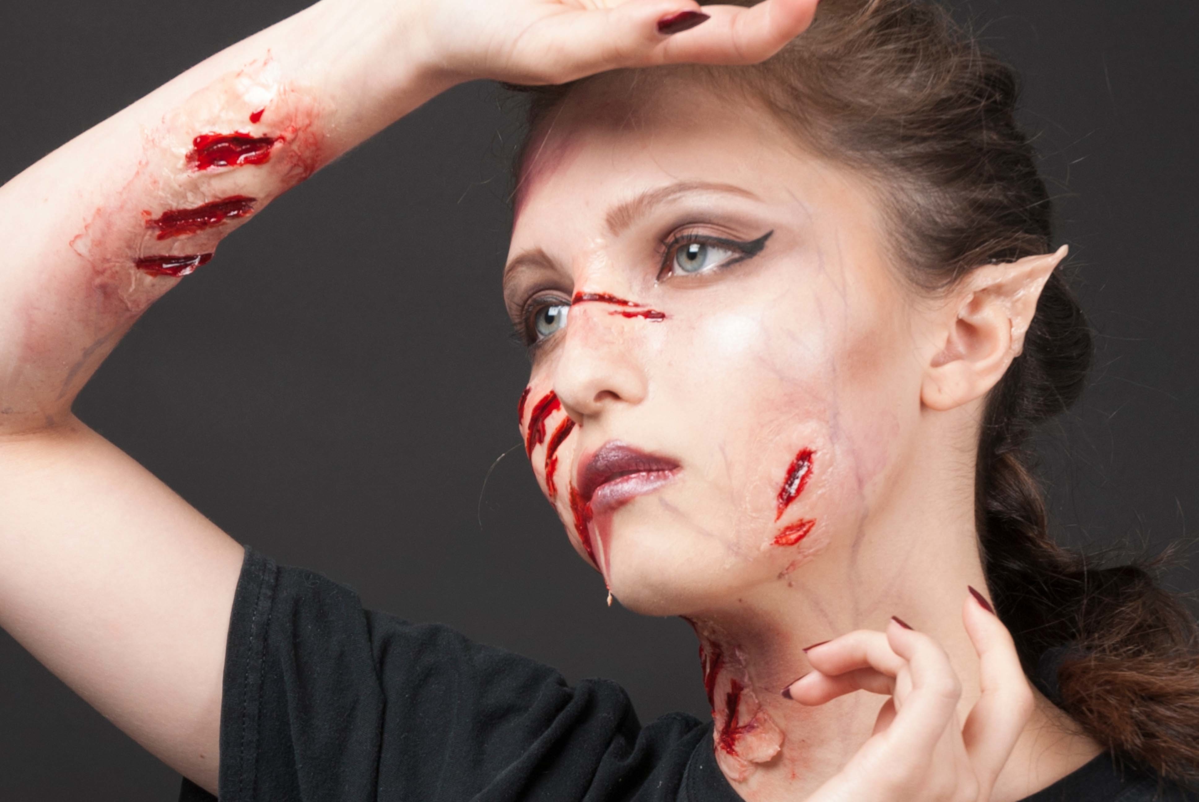 Headshot of a person with prosthetic wounds on their face, neck and wrist. They are also wearing prosthetic points on their ears.