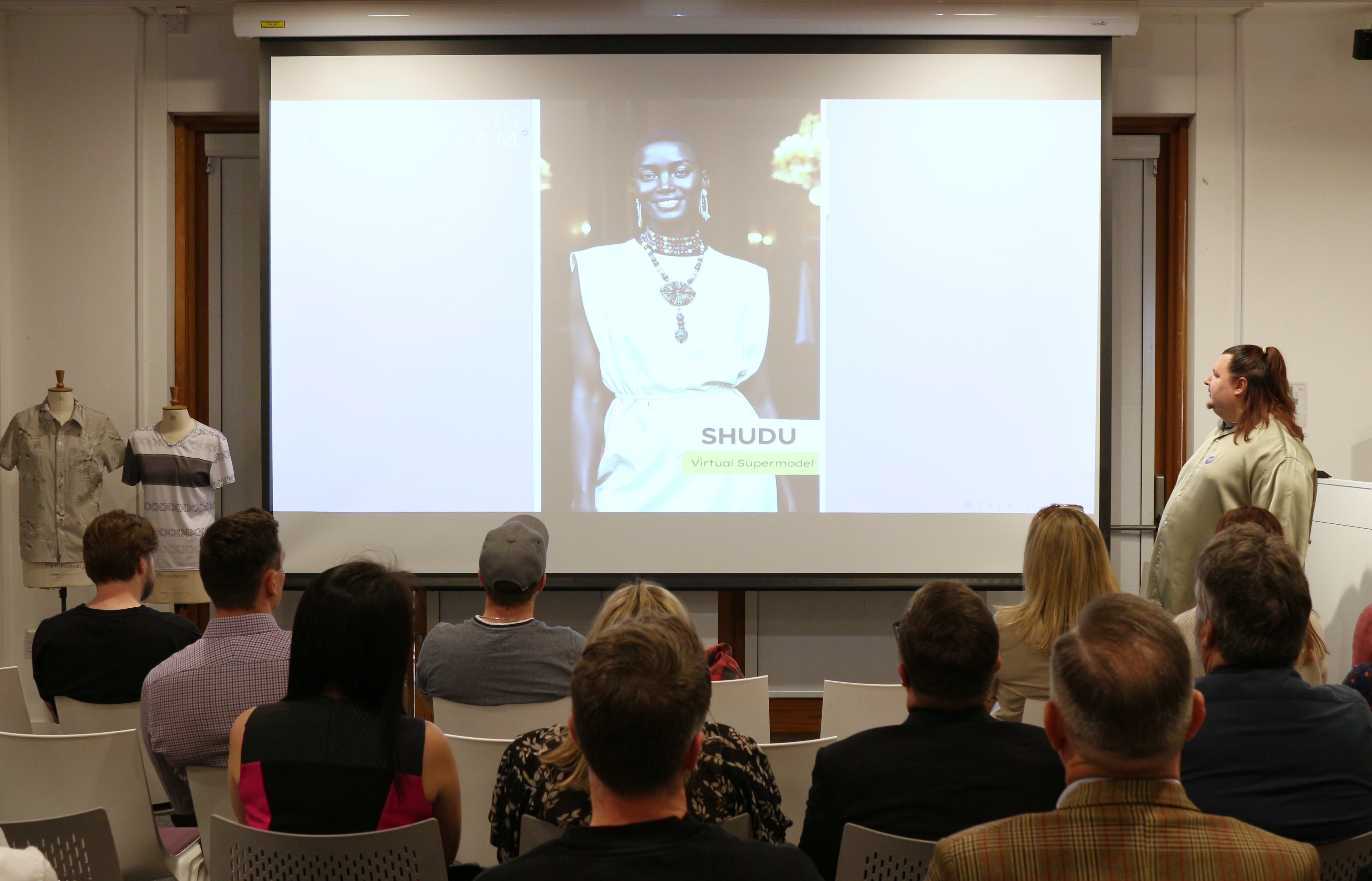 A large screen showing a presentation slide with an image of a virtual supermodel on it. An audience, viewed from the back, listen to the presentation.