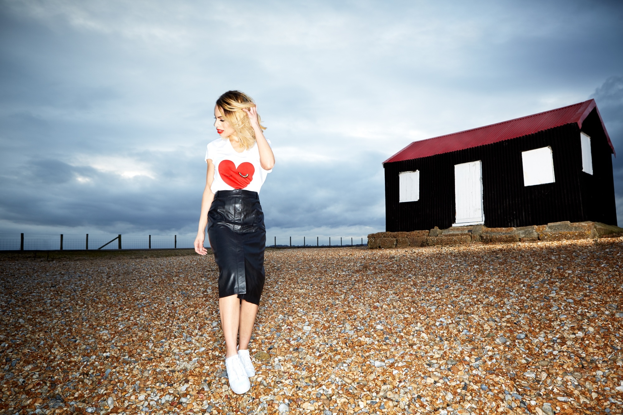 Woman in white heart t-shirt and black leather skirt walking on pebble beach towards black building with red roof under cloudy sky.
