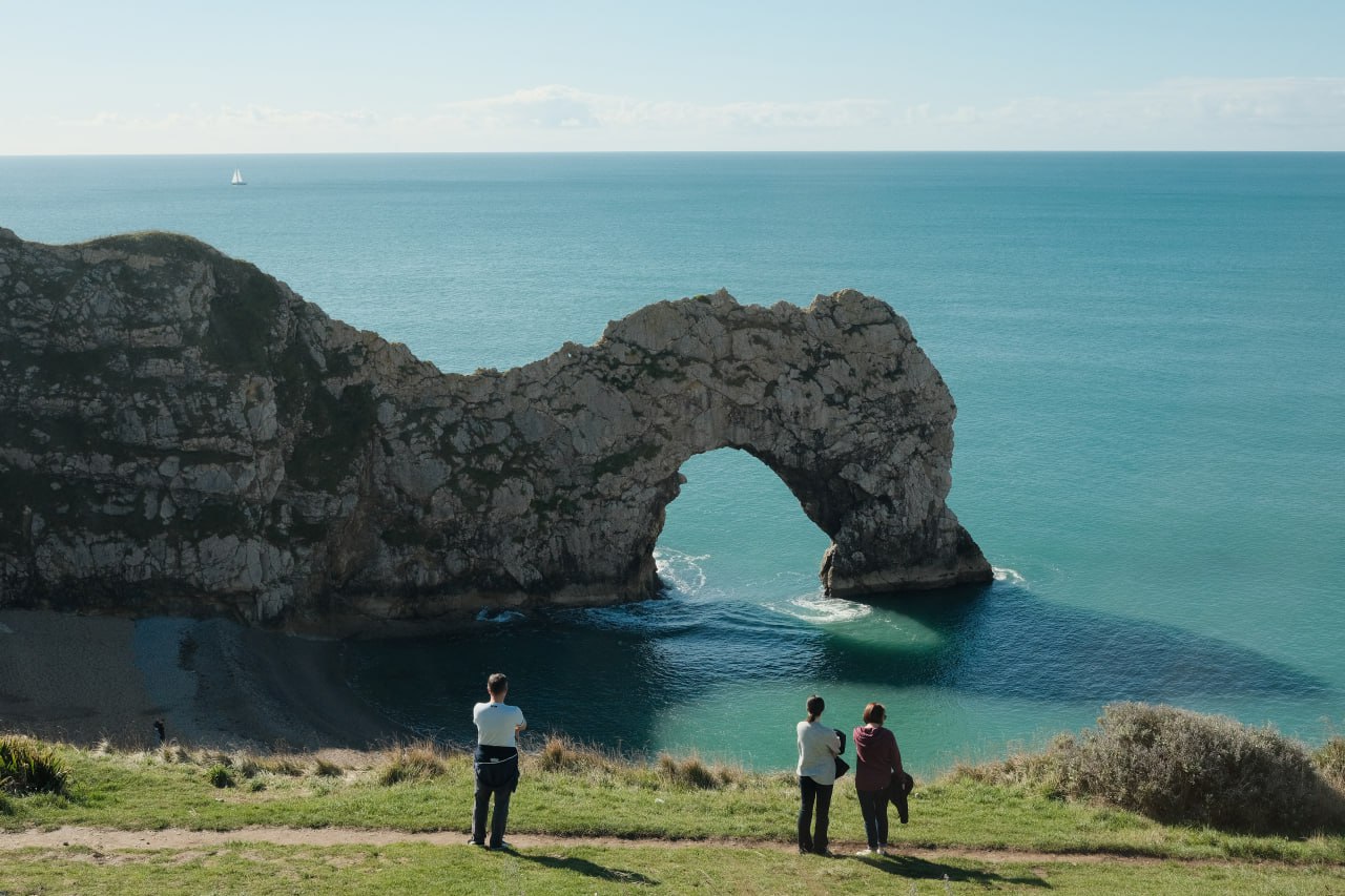 Three people stand on a grassy cliff overlooking the sea, with a natural stone arch, called Durdle Door, in the background. The calm water reflects the arch, and the sky is partly cloudy.