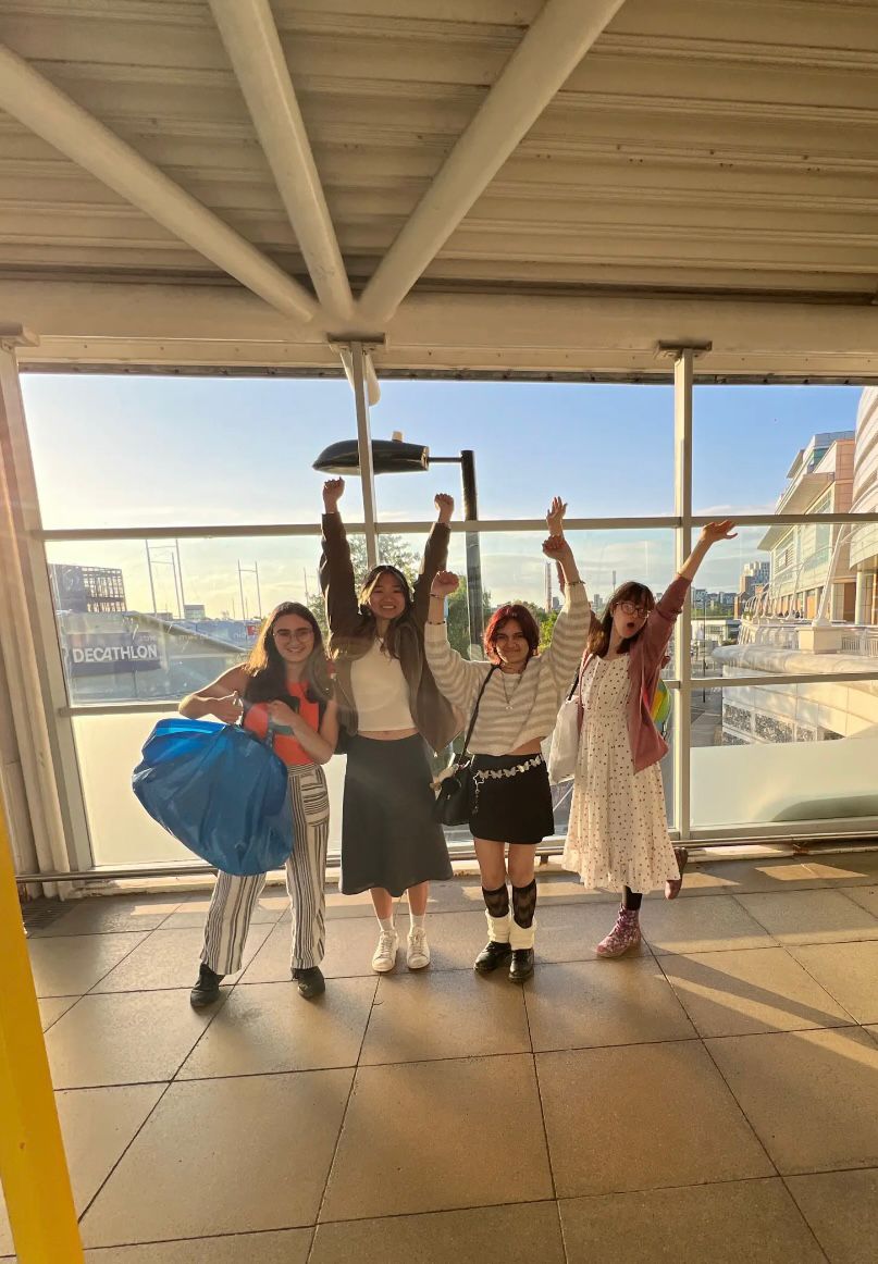 Four young people stand together at an airport terminal, posing and raising their arms. One woman holds a large blue bag, while the others wear various outfits including skirts and dresses. The background features large windows with sunlight illuminating the space.