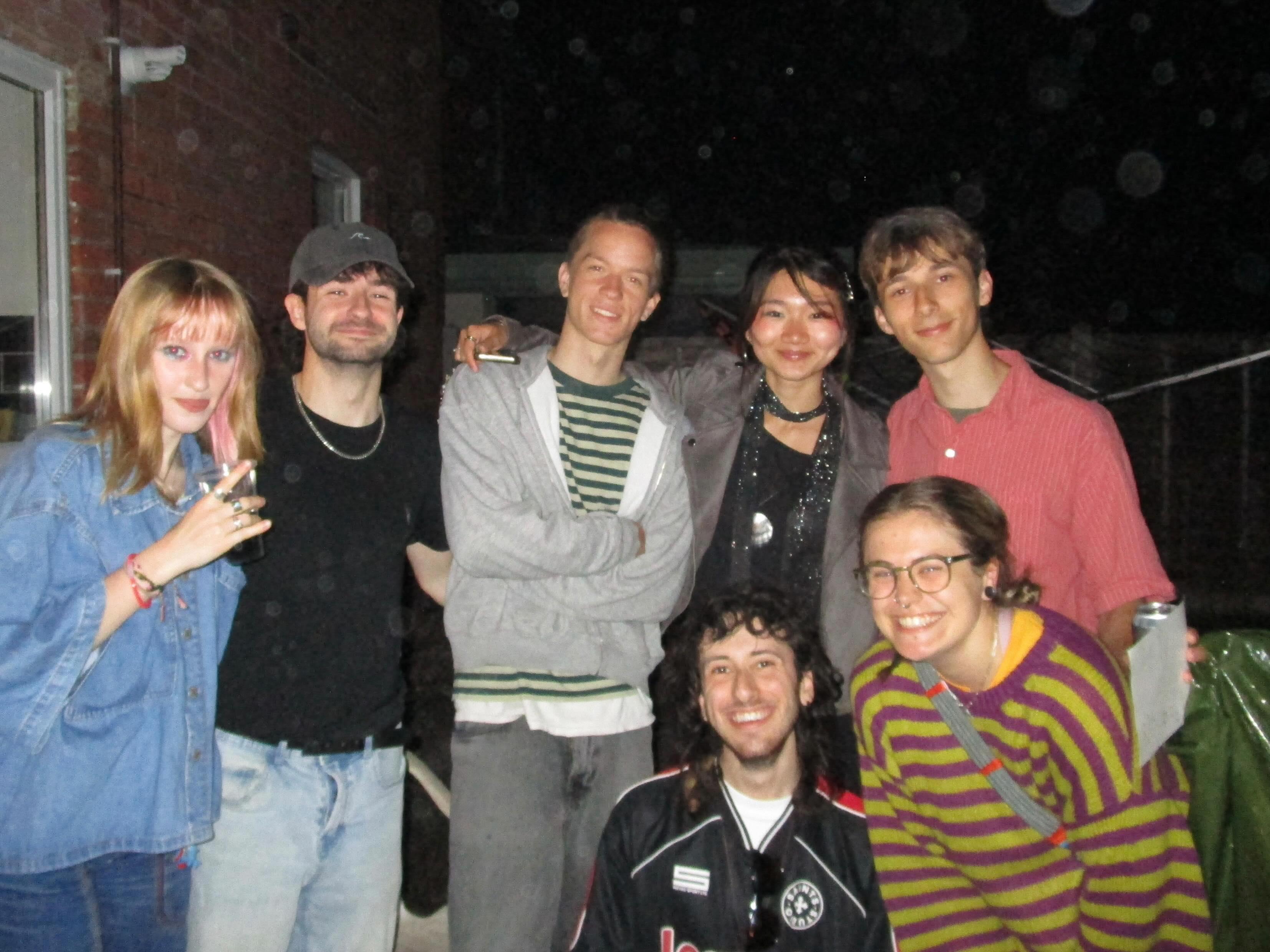 A group of seven people stands outdoors in the evening, with some wearing casual clothing. They are arranged closely together in front of a brick building, with a dark background, creating an informal gathering scene.