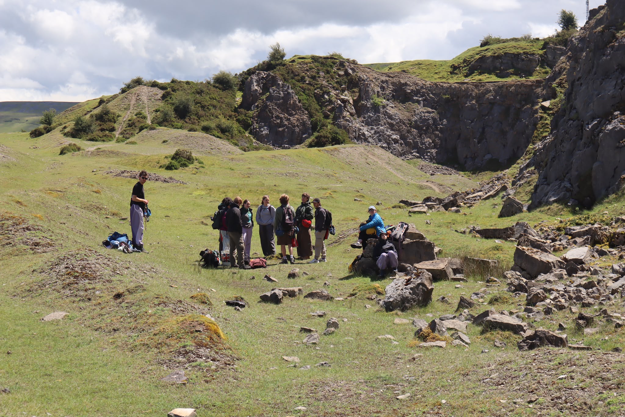 A group of eight people gathered on a grassy area surrounded by rocky terrain. They are standing and sitting next to backpacks, with hills and a cloudy sky in the background.