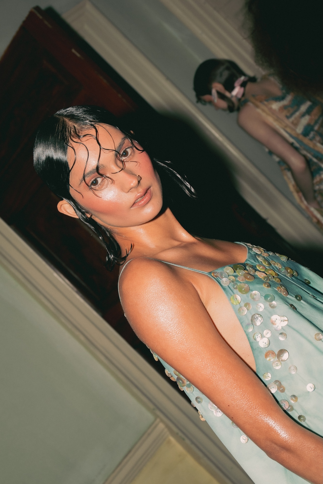 Woman with wet dark hair and glasses in bathtub wearing floral dress, another person visible in background