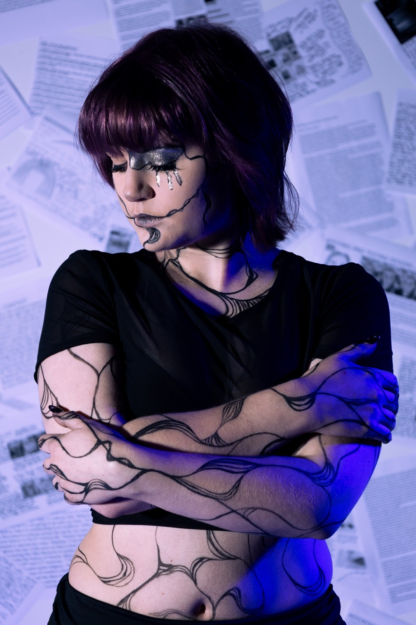 A young woman with short purple hair wears a black top and has intricate black body paint design on her arms. She stands with her arms crossed, and the background features scattered newspaper pages, illuminated with blue light.