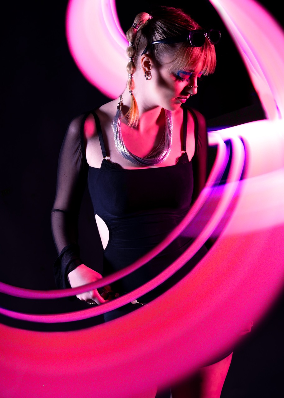 A woman wearing a black sleeveless top is standing in a dark space, surrounded by swirling pink and purple light patterns. She has a braid hairstyle and is wearing a silver necklace, while sunglasses rest on her head.