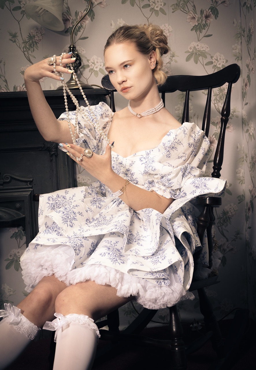 A young woman wearing a white dress with blue floral patterns sits in a wooden chair. She holds a string of pearls in one hand, adorned with various rings and jewellery, with a vintage wallpaper backdrop featuring floral motifs.