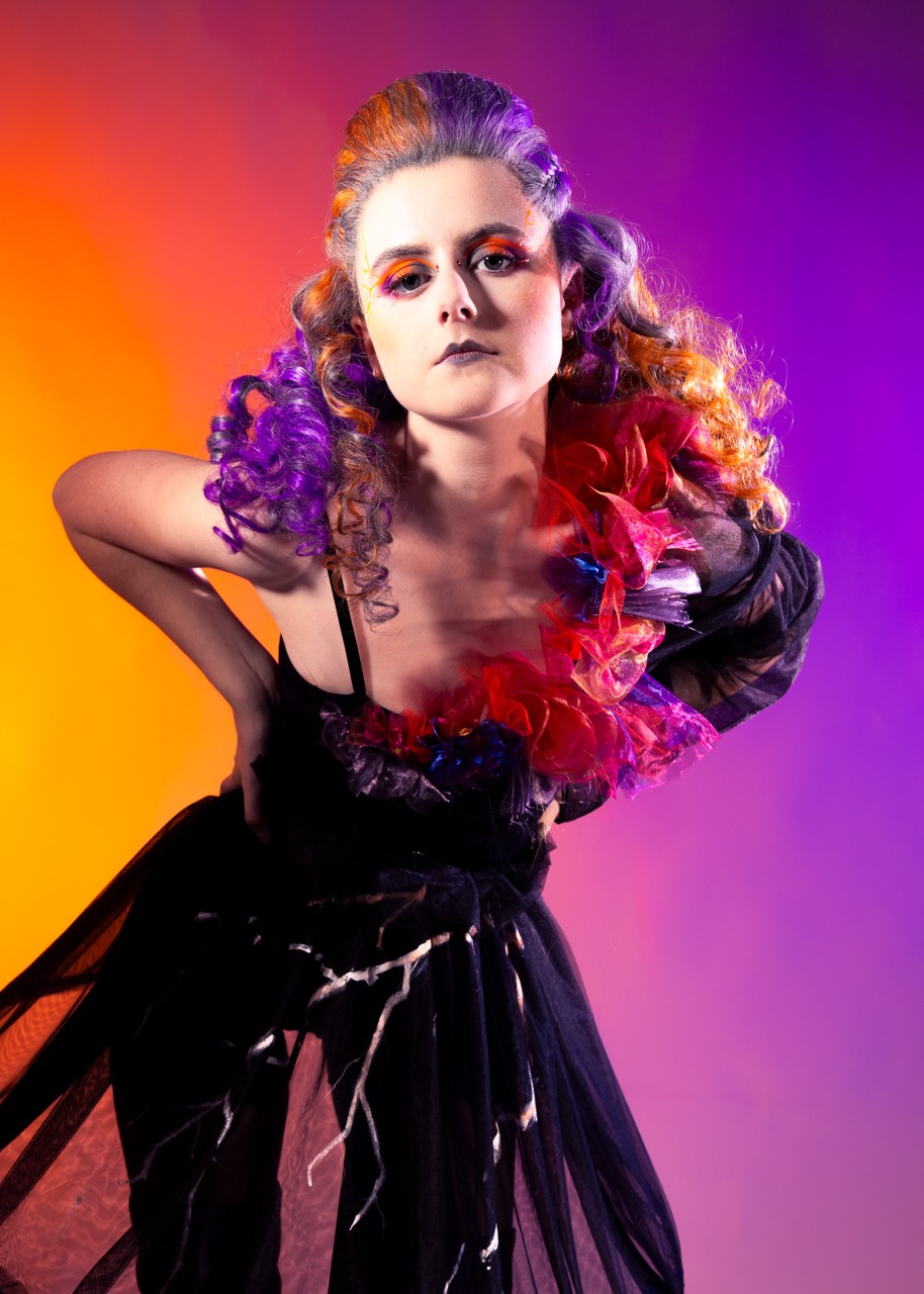 A model poses in a dramatic outfit, featuring a black dress and vibrant, multicoloured hair styled with curls. The background transitions from orange to purple, enhancing the bold look of the model and the vibrant flowers adorning her neckline.