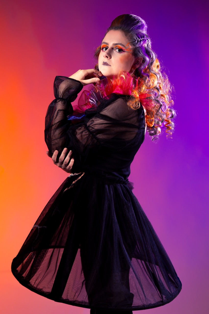 A model with curly, colourfully styled hair poses in a black sheer dress against a gradient background of orange and purple hues. The model's right hand is near her face, and she is looking towards the camera.