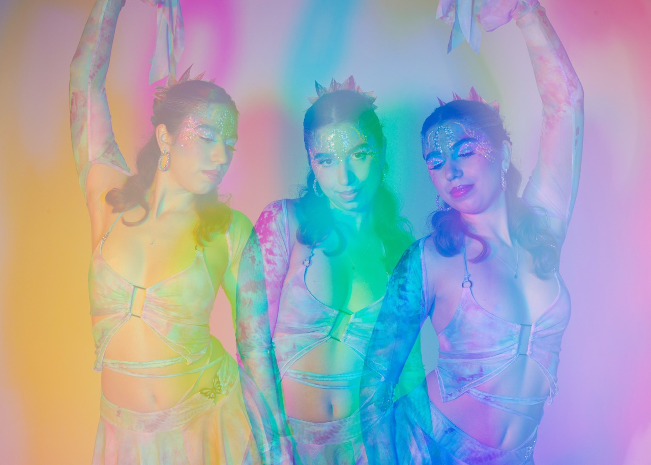 A woman dressed in a shimmering costume poses in three mirrored images, each surrounded by pastel rainbow colours. The costume features intricate designs and the woman is holding flowing scarves, creating a vibrant visual effect.
