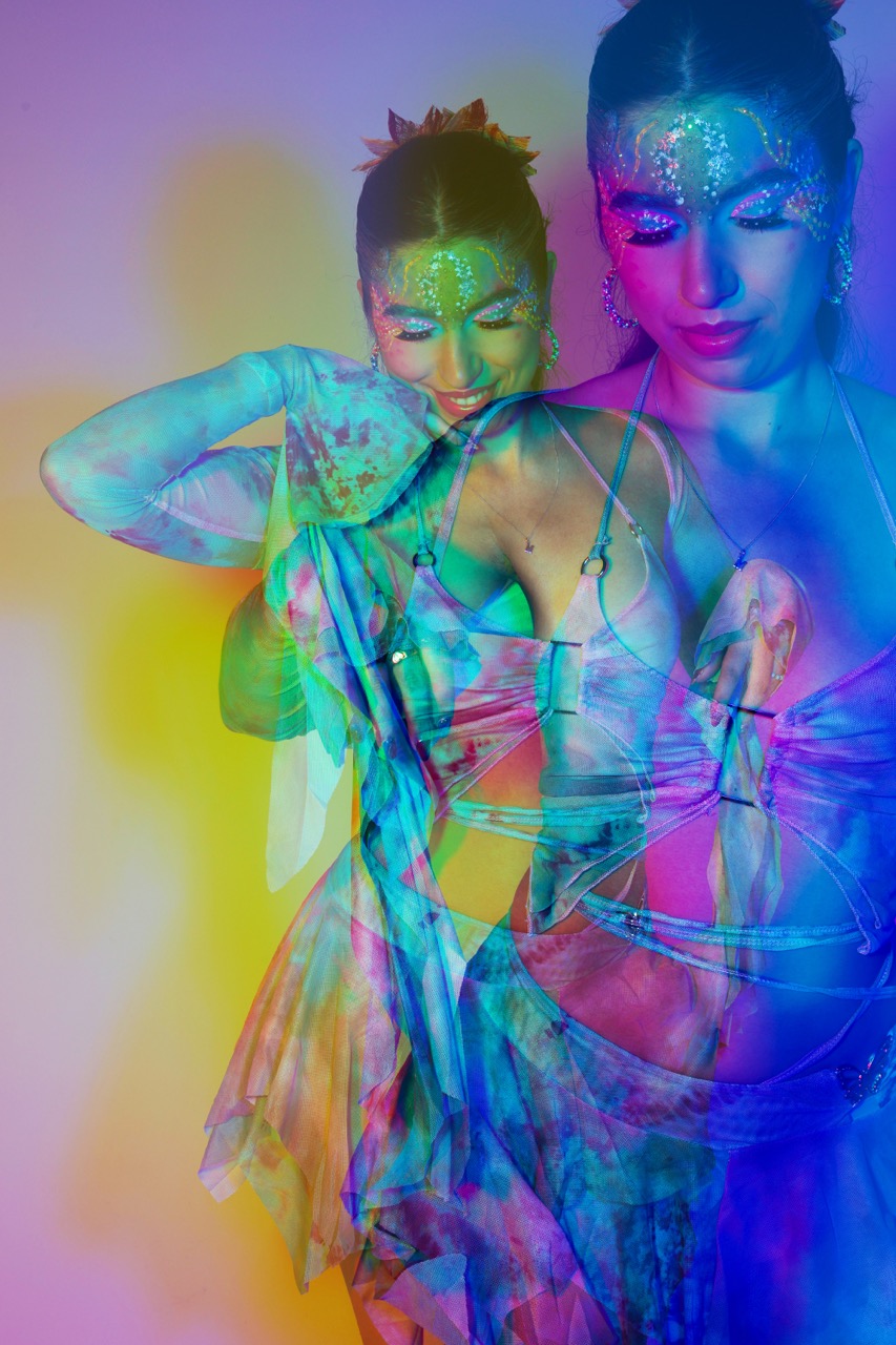A woman wearing a shimmering outfit with a colourful, layered design is shown against a vibrant gradient background. Her makeup features glitter and elaborate face paint, enhancing the visual effects of the image.