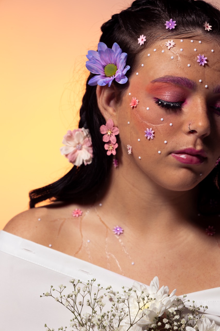 A young woman with wavy dark hair adorned with flowers and miniature floral decorations on her face. She wears muted pink makeup and poses with her eyes closed, holding a bouquet of white flowers against a warm orange background.
