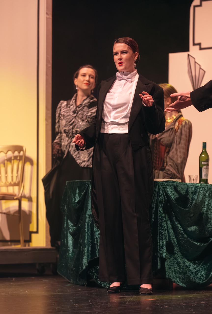 A performer in a black tuxedo with a white bow tie gestures while speaking on stage. In the background, two other actors are partially visible, one wearing a patterned outfit and the other seated near a table with drinks and a green tablecloth.