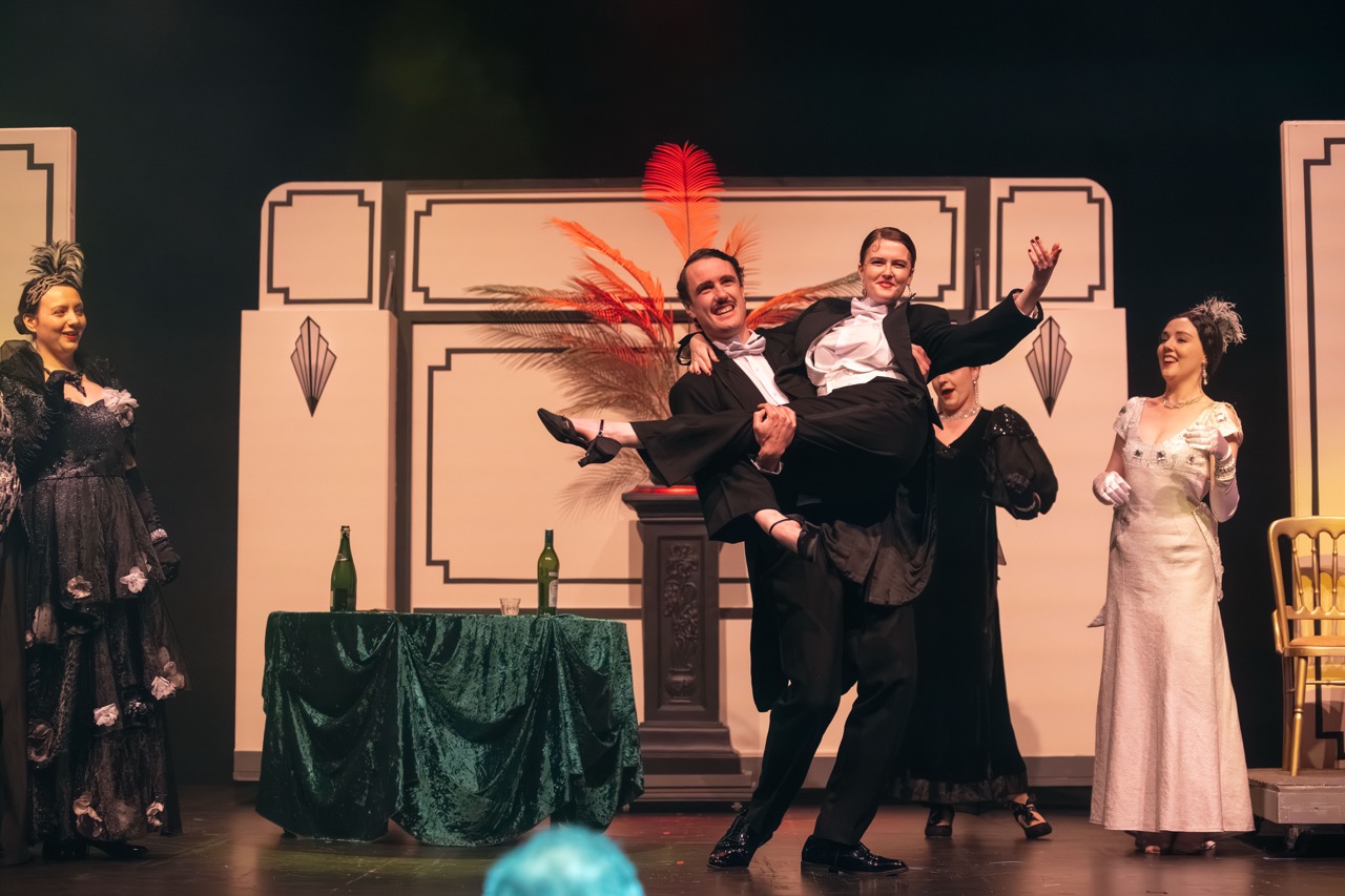 A man in a tuxedo lifts a woman in a white dress during a theatrical performance. They are surrounded by three other performers: two women in black dresses and one woman in a white gown, with a green table and a decorative backdrop behind them.