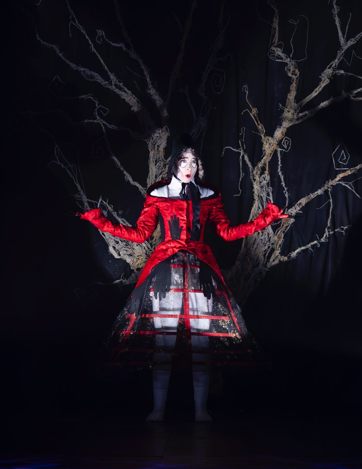 A performer dressed in a red and black theatrical costume stands against a dark backdrop featuring an illustration of a tree. The costume has a fitted red bodice and a sheer black skirt, and the performer extends their arms outward.