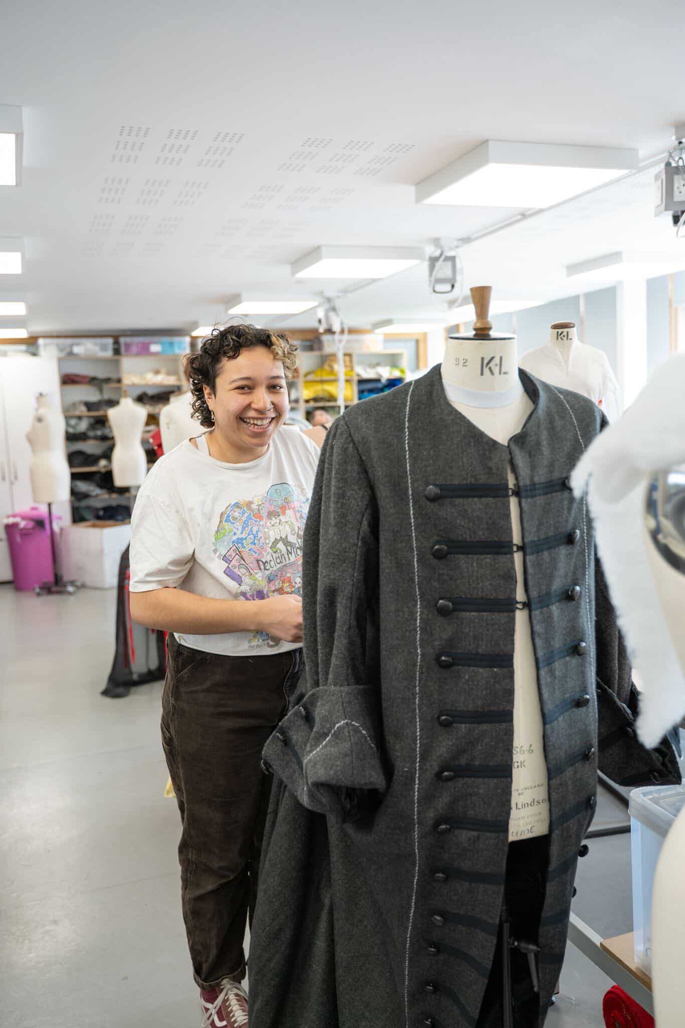 A woman with curly hair stands in a garment workshop, smiling as she adjusts a grey coat on a dress form. Behind her, various fabrics and mannequins are visible in a well-lit room with white walls.