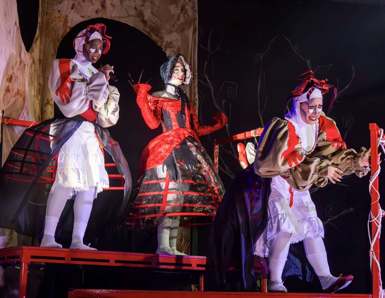 Three performers on a stage wearing elaborate costumes. The central performer is dressed in a red and black gown with a dramatic headdress, while the others are in contrasting white and beige outfits. The background features a dark, abstract set design.