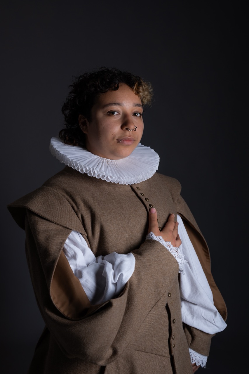 A person with curly hair wearing a large white ruff collar and a brown outer garment with wide sleeves. The figure is standing against a dark background, with one hand placed over the chest.