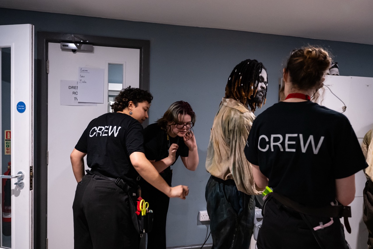 A group of three crew members in black shirts with 'CREW' printed on the back, interacting in a backstage area. One person stands near a life-sized mannequin with painted features, while another talks to a third crew member.