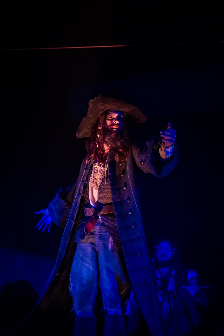 A performer dressed as a pirate stands on stage, wearing a black hat and a long coat. He has long brown hair and holds a weapon, with blue lighting creating a dramatic effect in the background, where other actors are slightly visible.